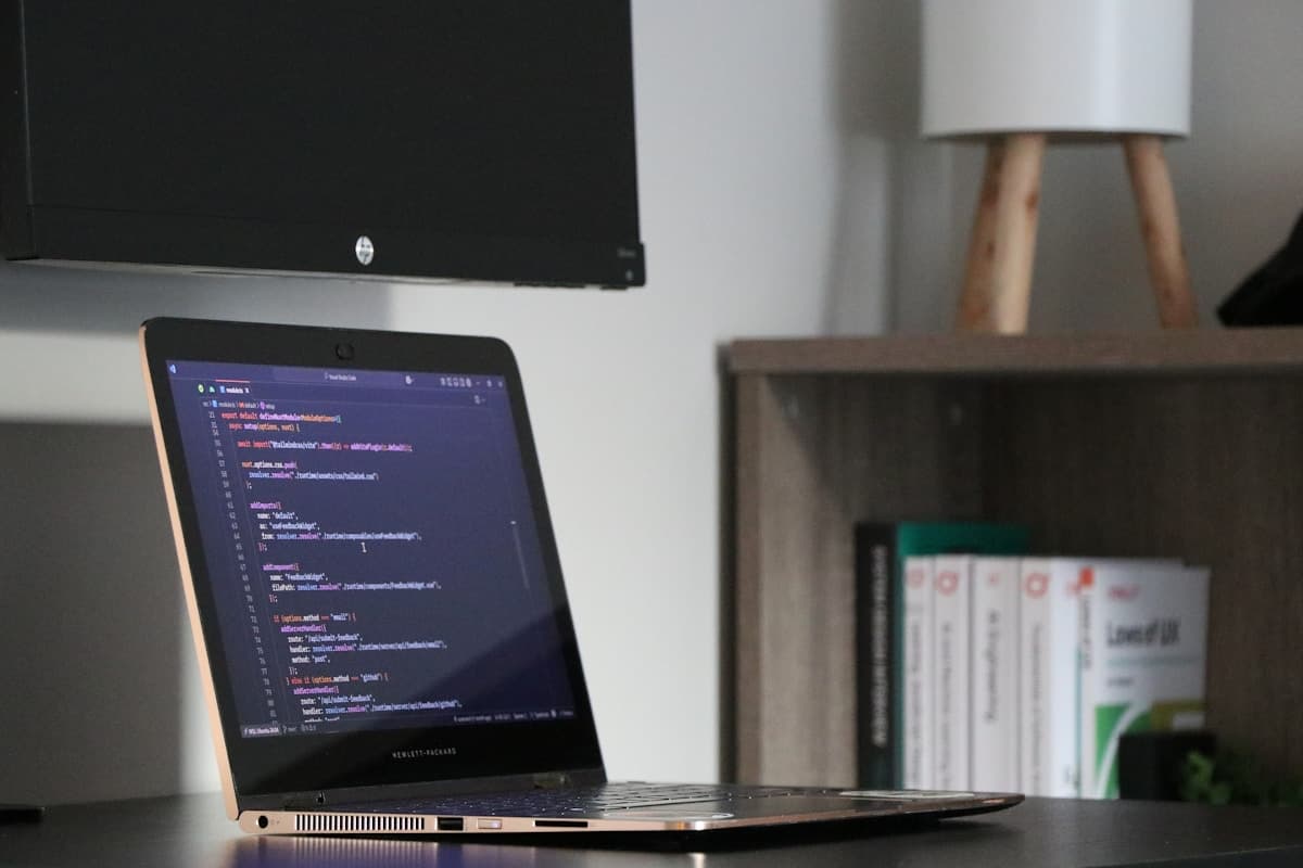Laptop displaying code on a desk with monitor and bookshelf
